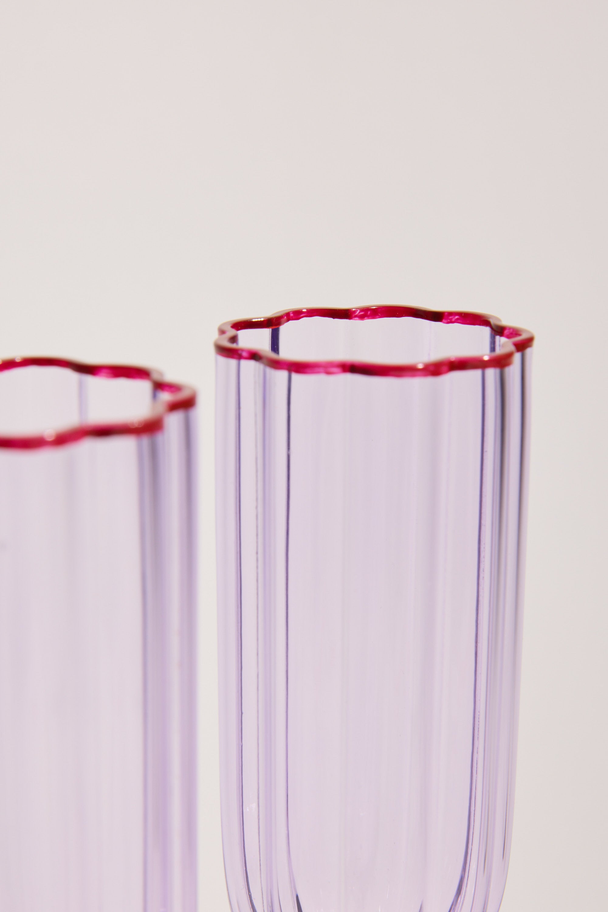 Two translucent purple Homelab Petal Champagne Flutes with red-outlined scalloped rims, from the Homelab brand, are shown against a plain light background. Only the upper parts of this elegant glassware set are visible.