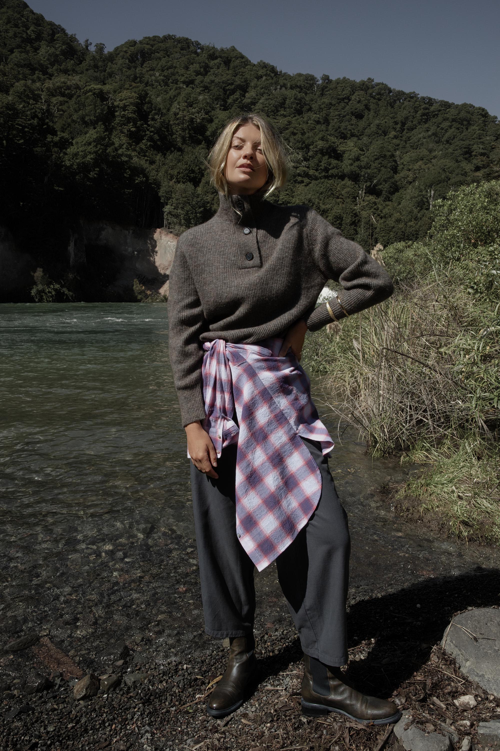 Woman by a river in the Elementary Collar Detail Sweater, forested hills behind her.