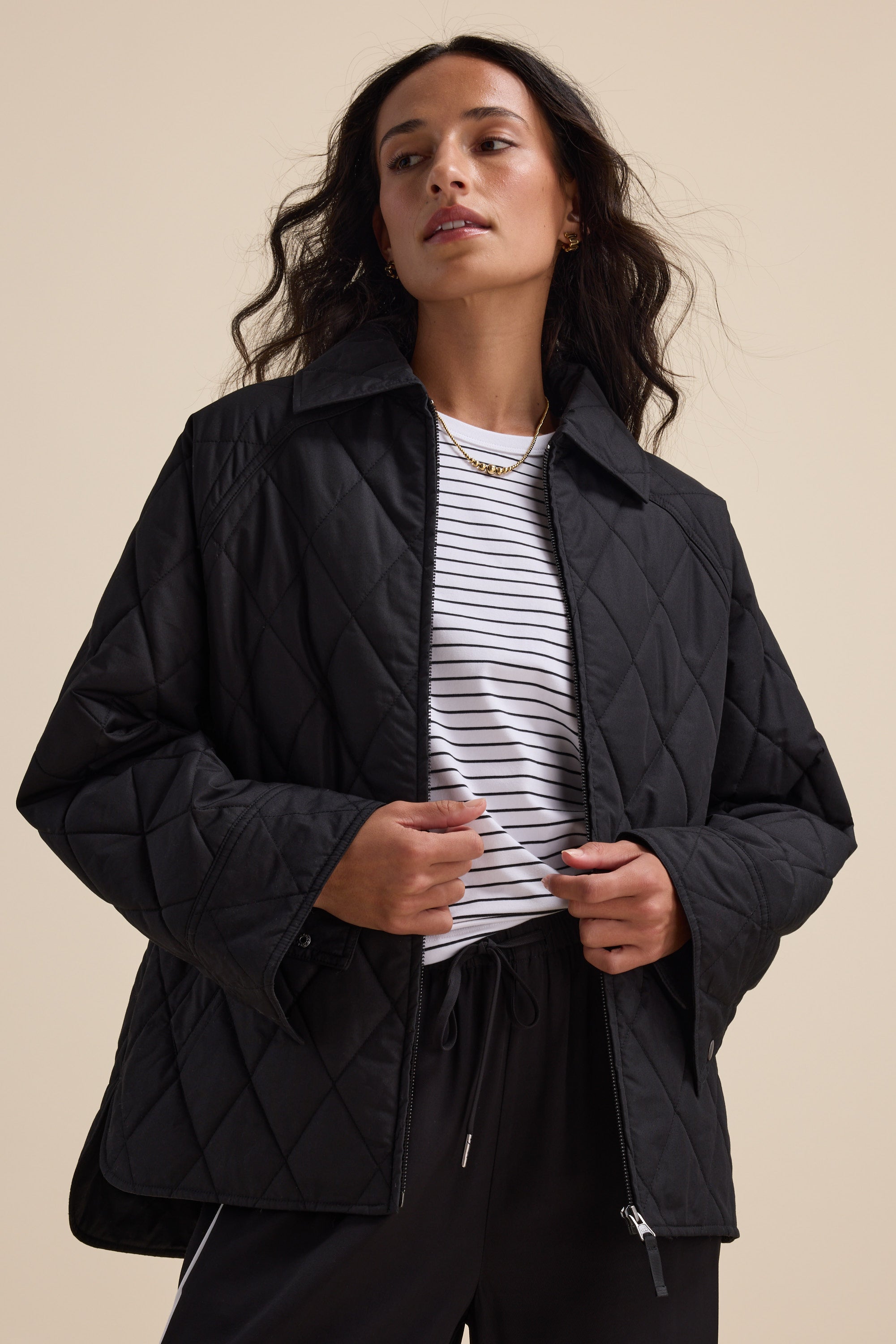 Woman in Elementary Quilted Puffer over striped shirt and black pants against beige background.