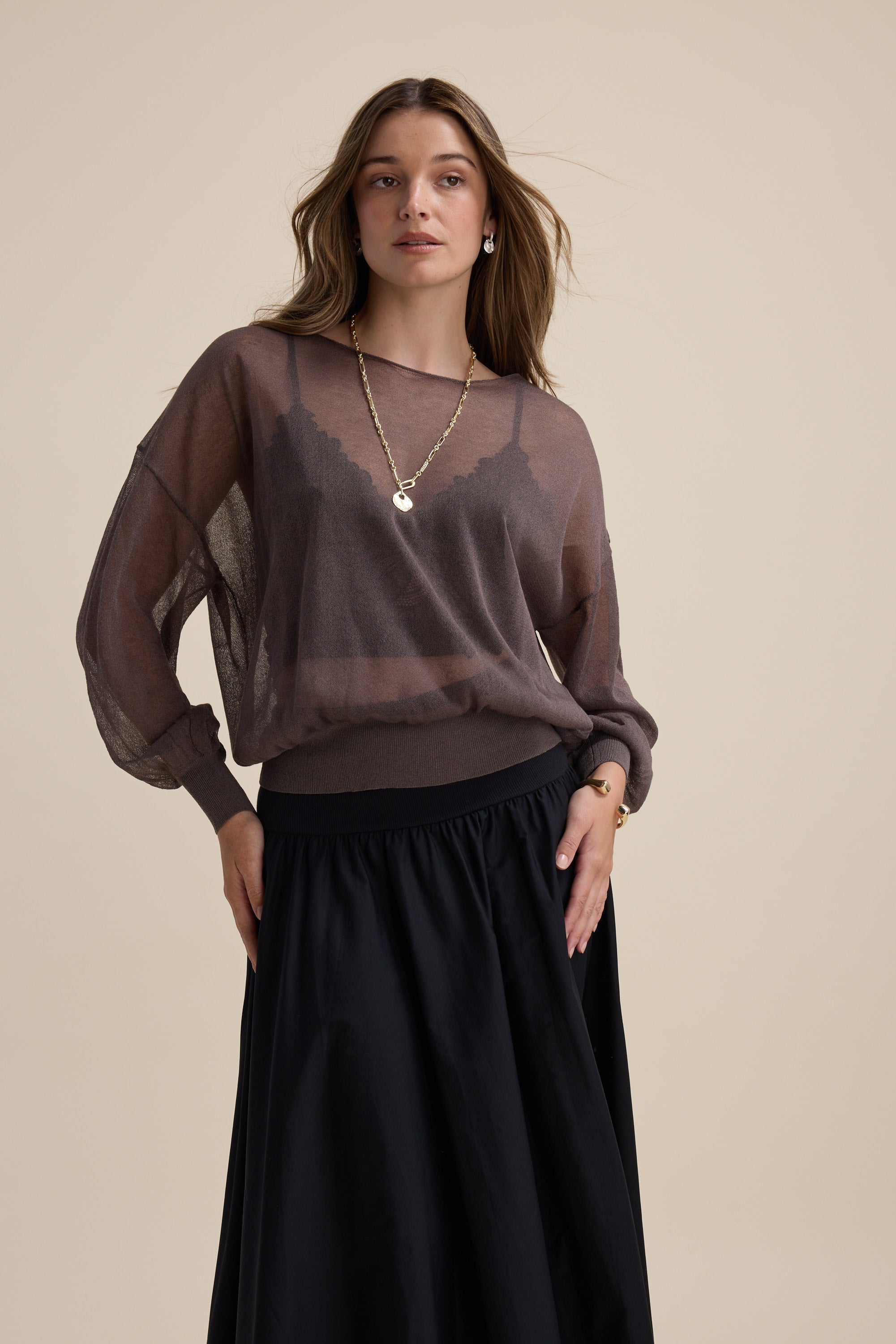 Woman wears Max Addie Sheer Knitted Top, black skirt, and gold necklace against beige background.