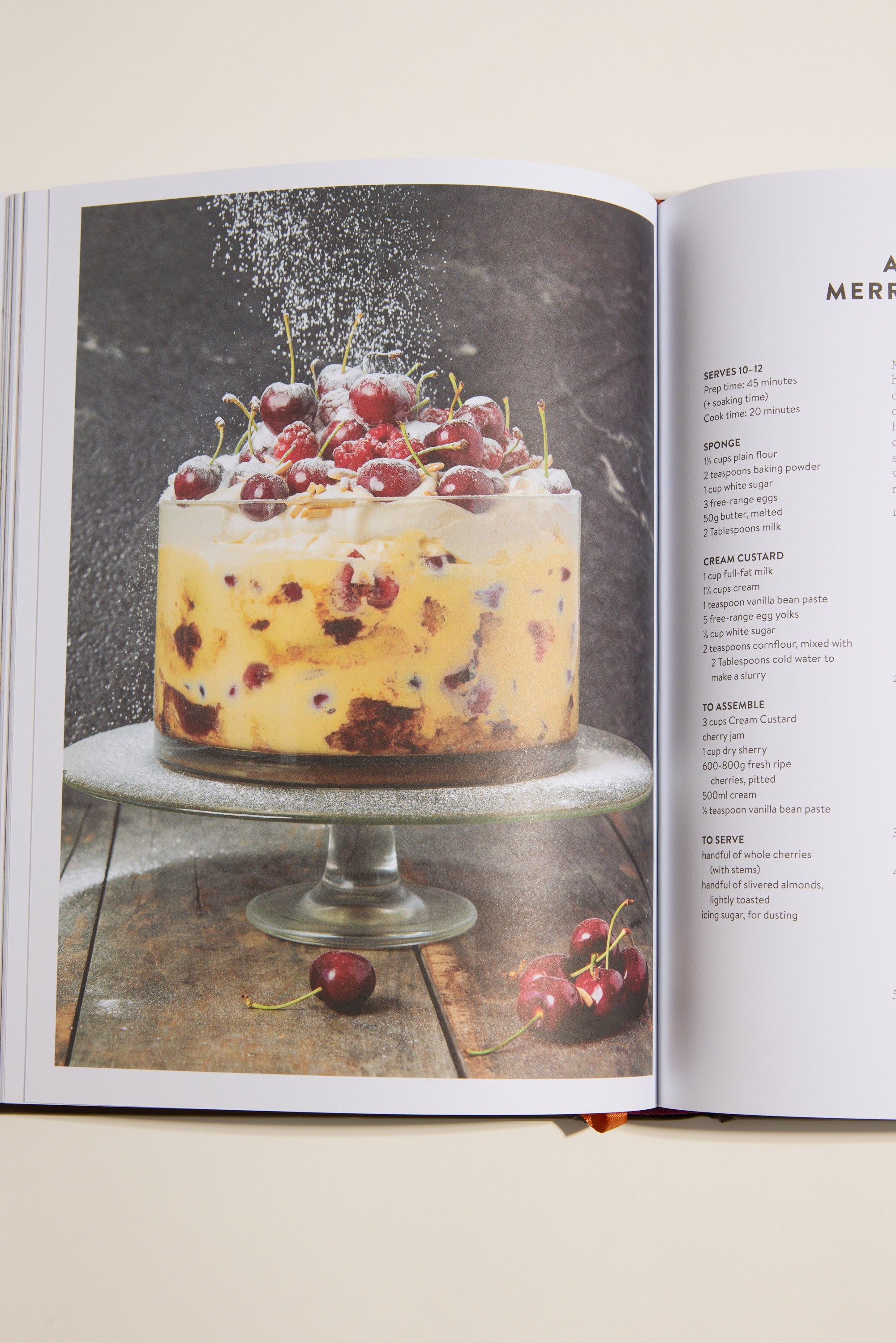 A trifle dessert in a glass bowl with whipped cream and cherries—just like a recipe from Nadia's Farm Kitchen Book by Other—sits on a cake stand as powdered sugar falls and cherries are scattered on the wooden surface below.