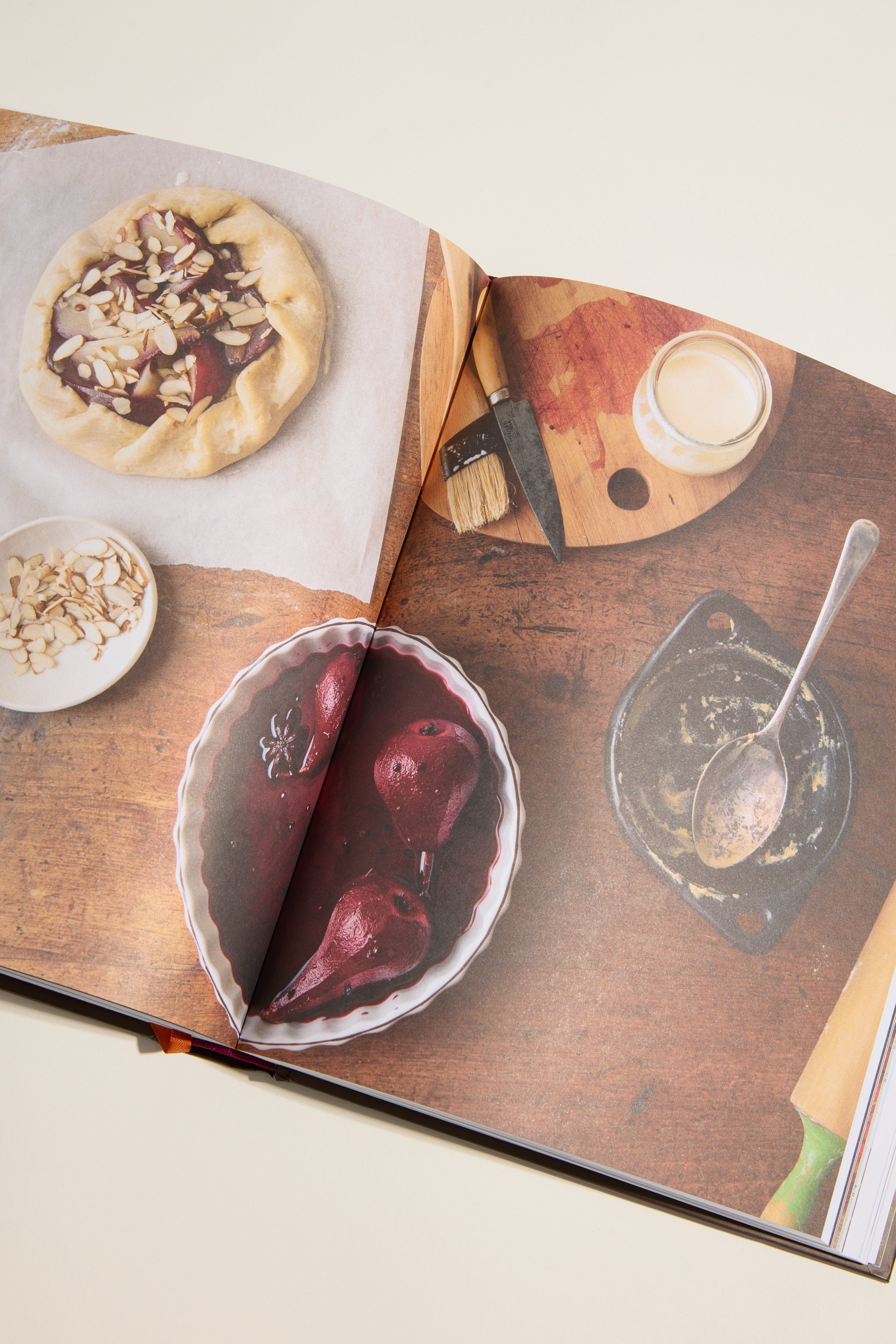 An open Nadia's Farm Kitchen Book by Other rests on a rustic wooden table with a fruit galette, poached pears in syrup, sliced almonds, a pastry brush, and baking utensils—ideal for trying seasonal recipes.