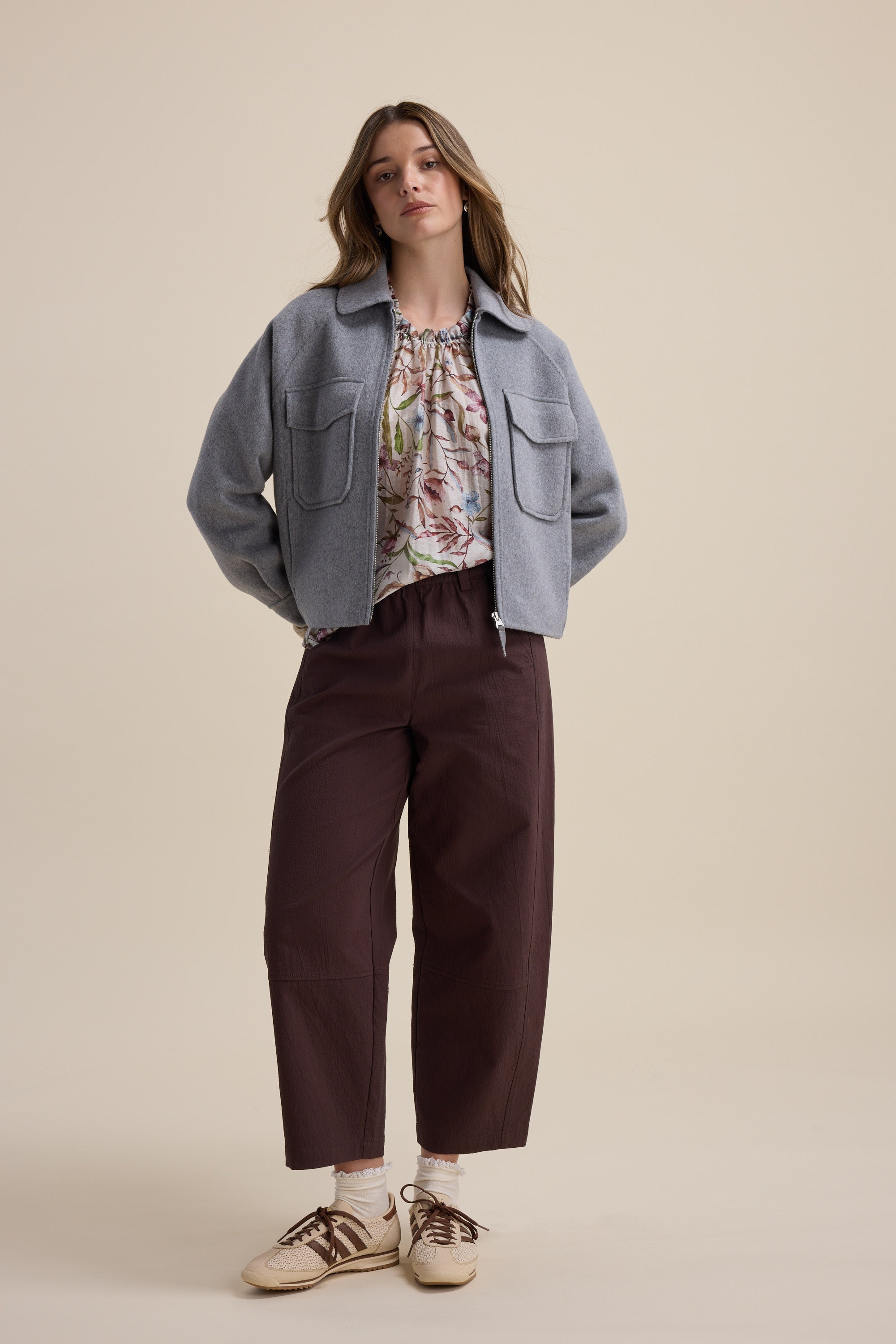 Woman in a gray jacket, Max Quinn Gather Detail Blouse, brown pants, and sneakers stands on beige.