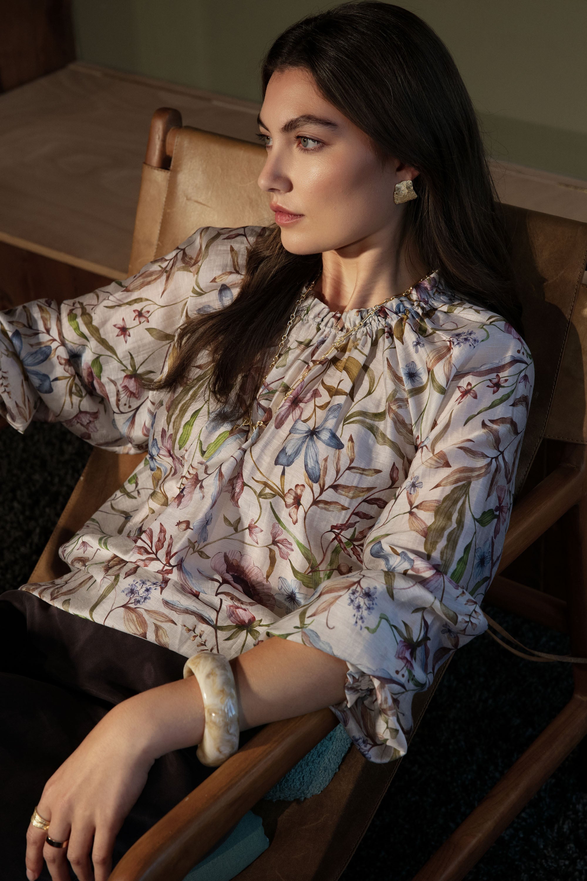 Woman in Max's Quinn Gather Detail Blouse sits on a wooden chair, looking thoughtfully aside.