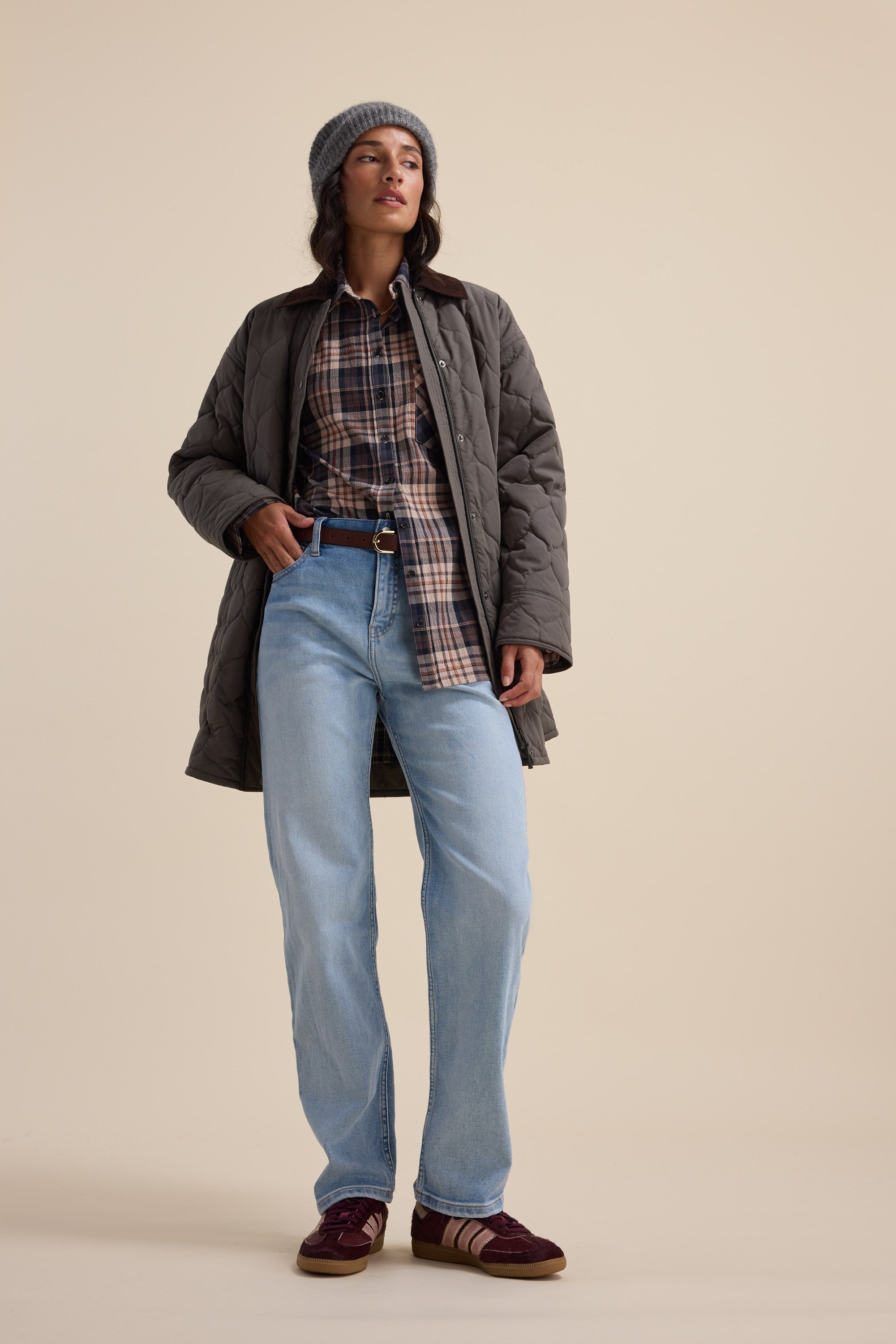 Woman in Elementary Textured Shirt, jeans, coat, beanie & sneakers on beige backdrop.
