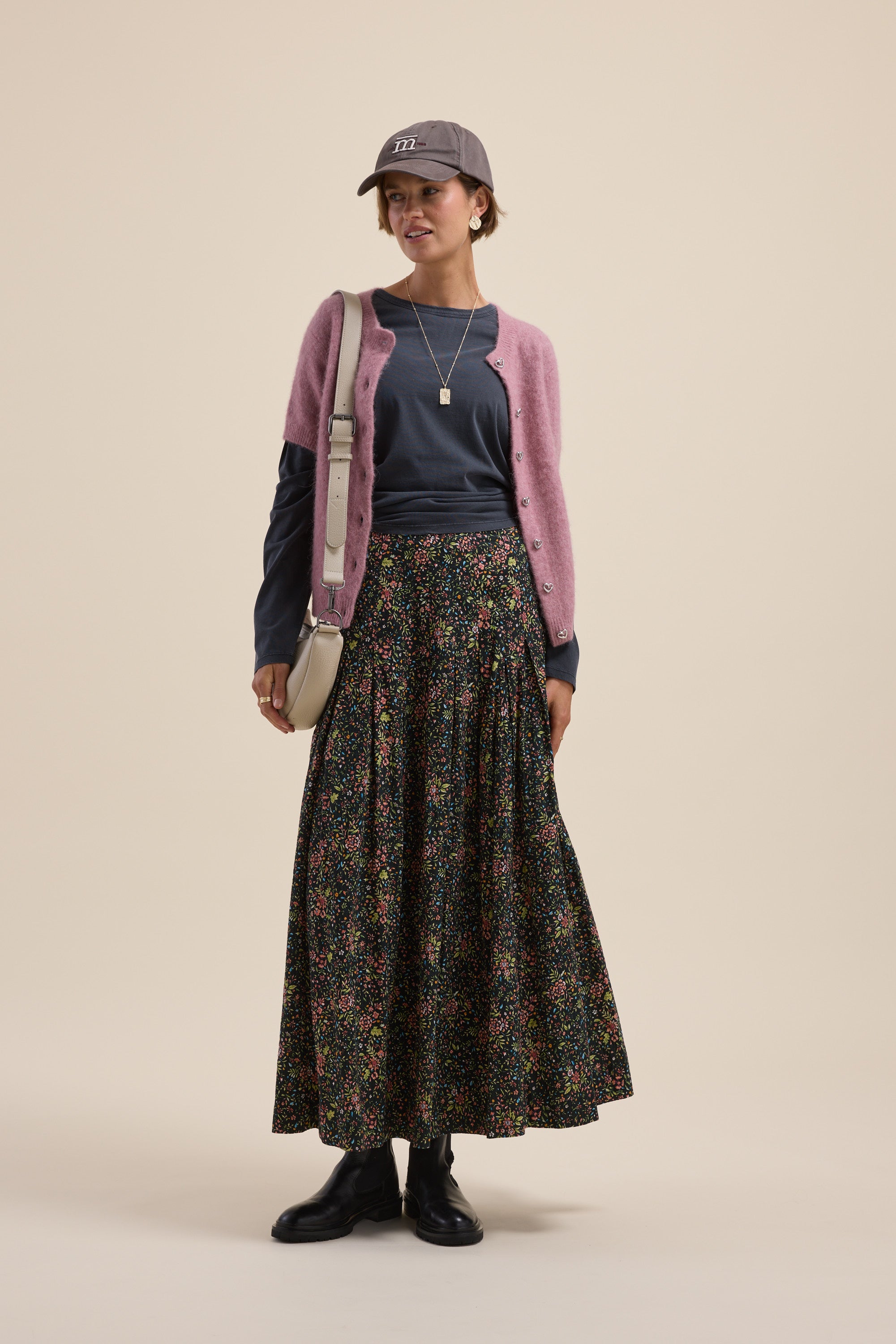 Woman in a cap, pink cardigan, Max Elementary Slouchy Long Sleeve Tee, floral skirt, boots, and crossbody bag.