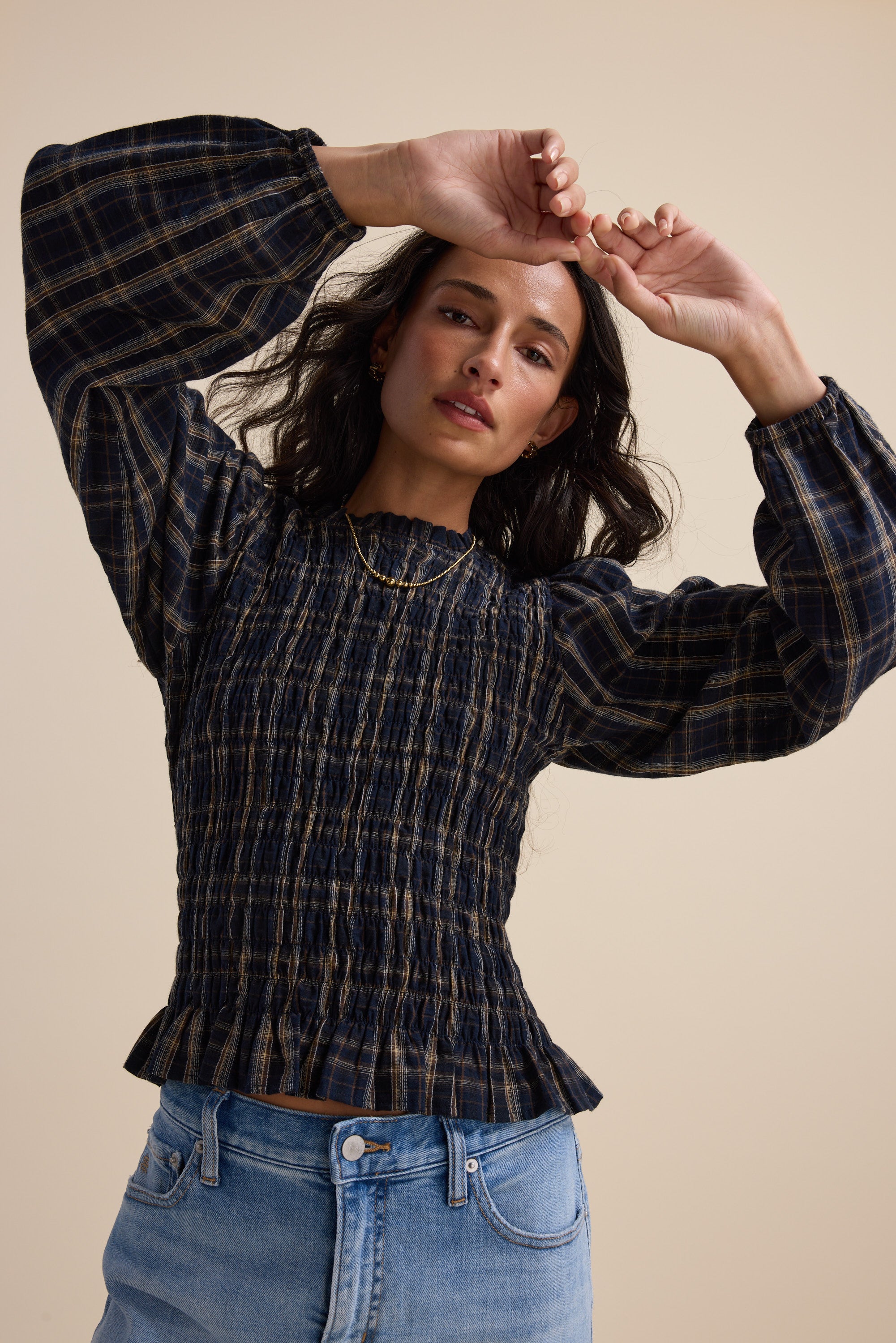 Woman in an Elementary Shirred Long Sleeve Top and jeans poses with arms raised against beige.