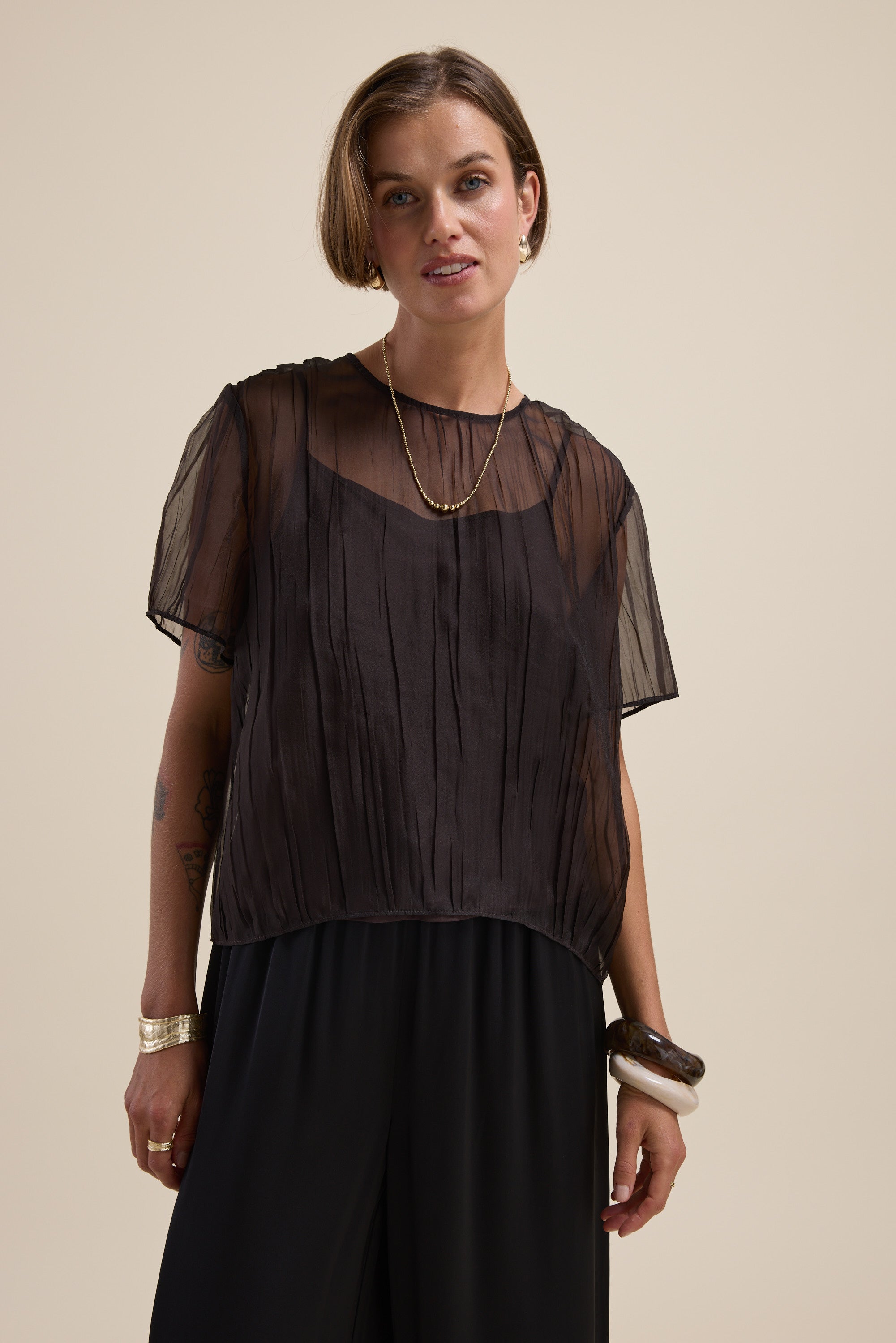 Model wears the Cass Sheer Top by Max with black pants and layered necklaces on a beige background.