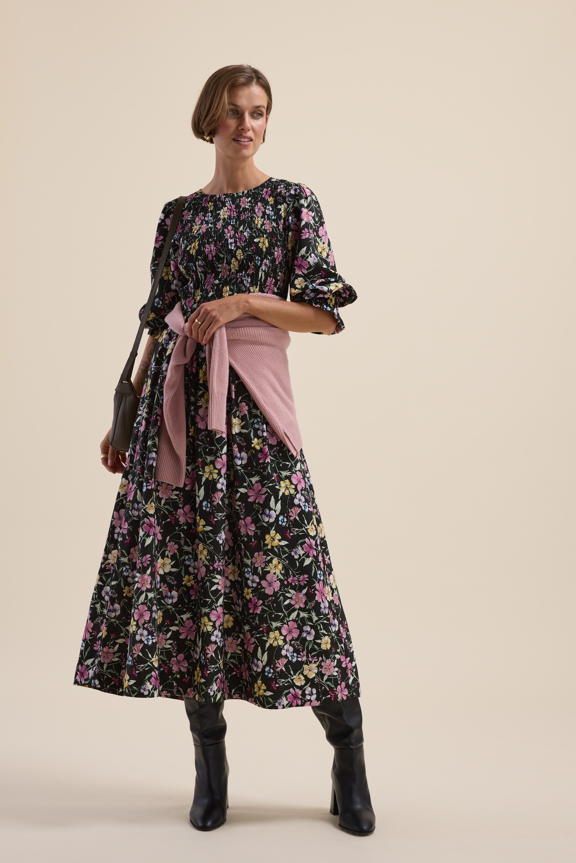 Woman in a Max Brooke Shirred Long Sleeve Dress, pink sweater at waist, black boots, holding a bag.