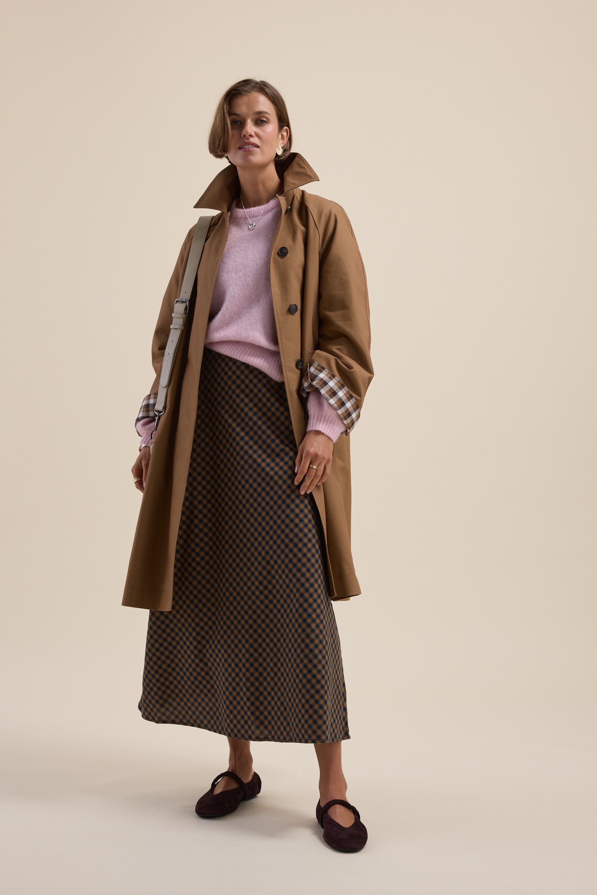 Woman in the Max Lucy Bias Cut Dress, tan trench coat, pink sweater, and dark flats standing.