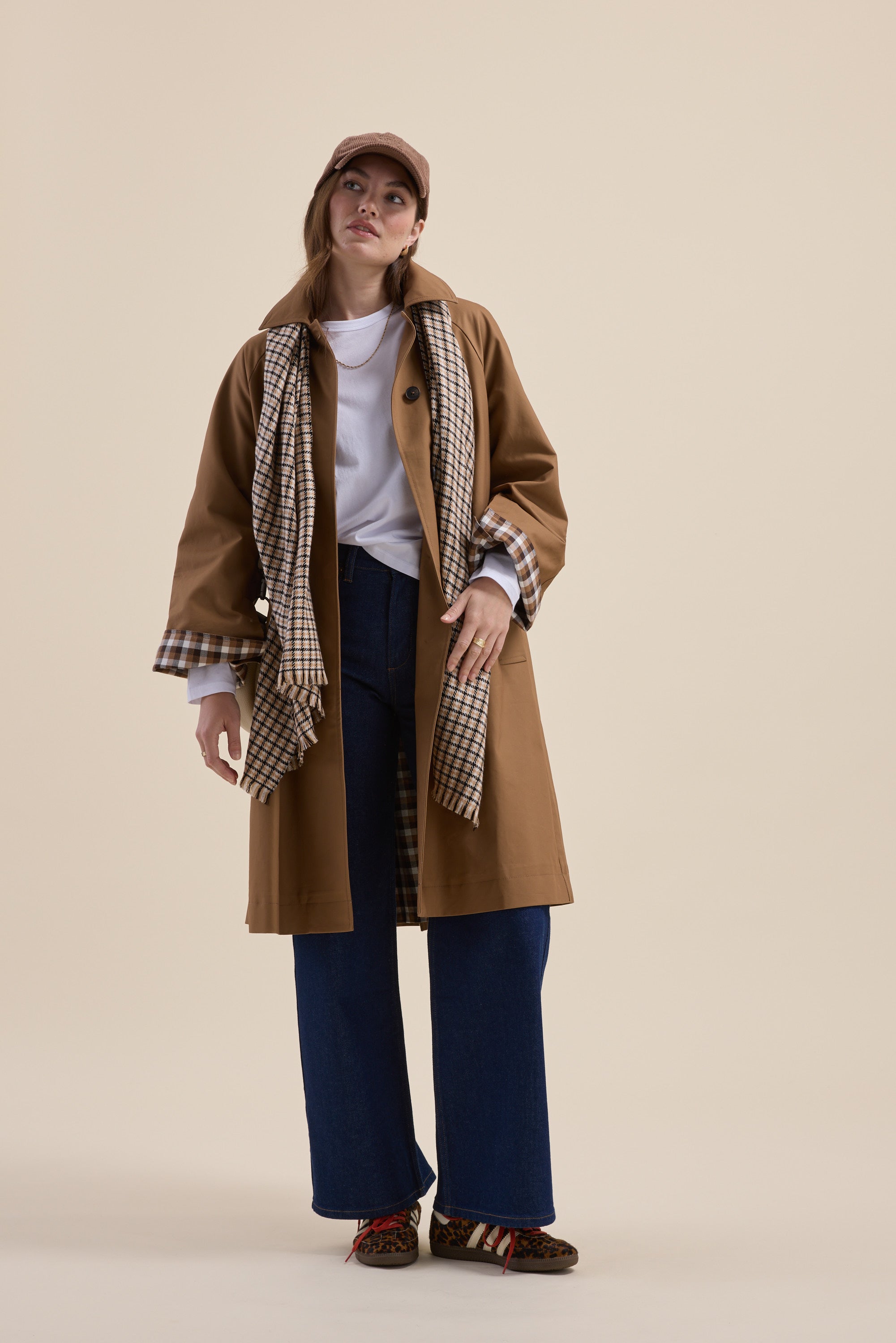 Woman in a Max London Trench Coat with plaid scarf, wide-leg jeans, and cap on beige background.