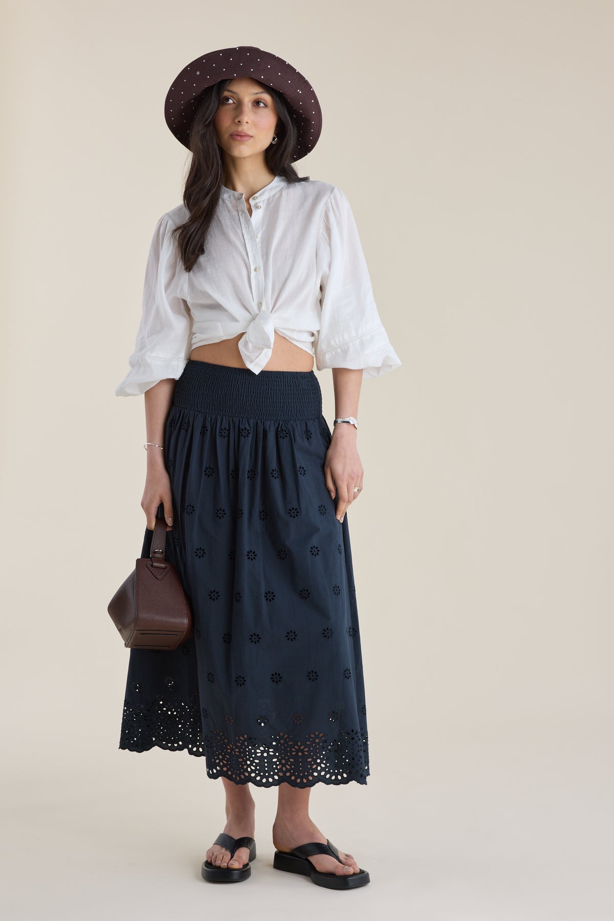 Woman in Elementary broderie skirt, white blouse, brown hat, holding bag, and sandals.