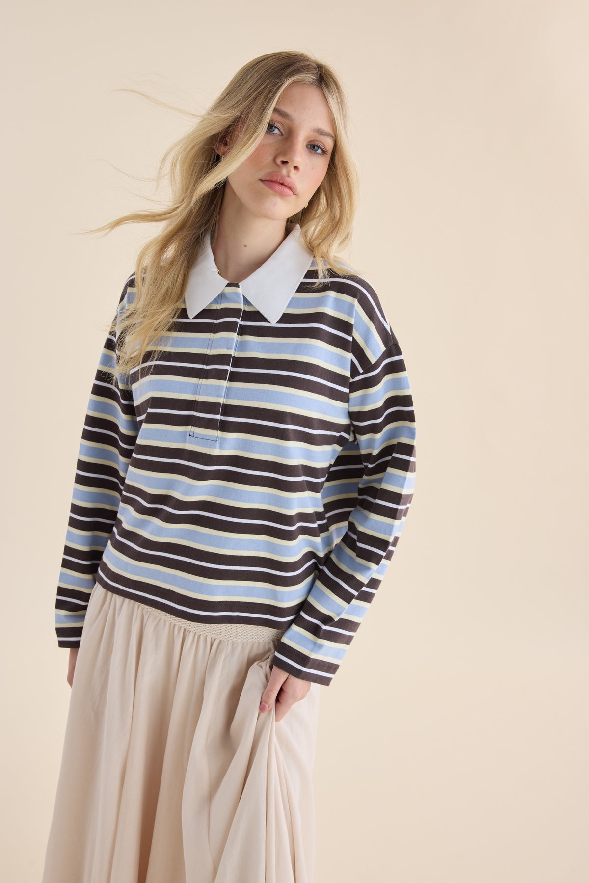 Woman in Elementary Ice Blue Stripe Polo Top and cream skirt, posing by beige background.