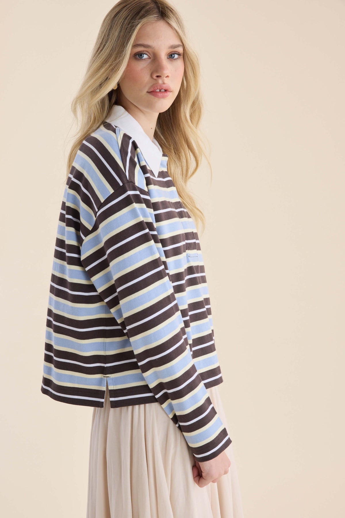 Woman in an Elementary Polo Top, Ice Blue Stripe, stands sideways against a beige backdrop.