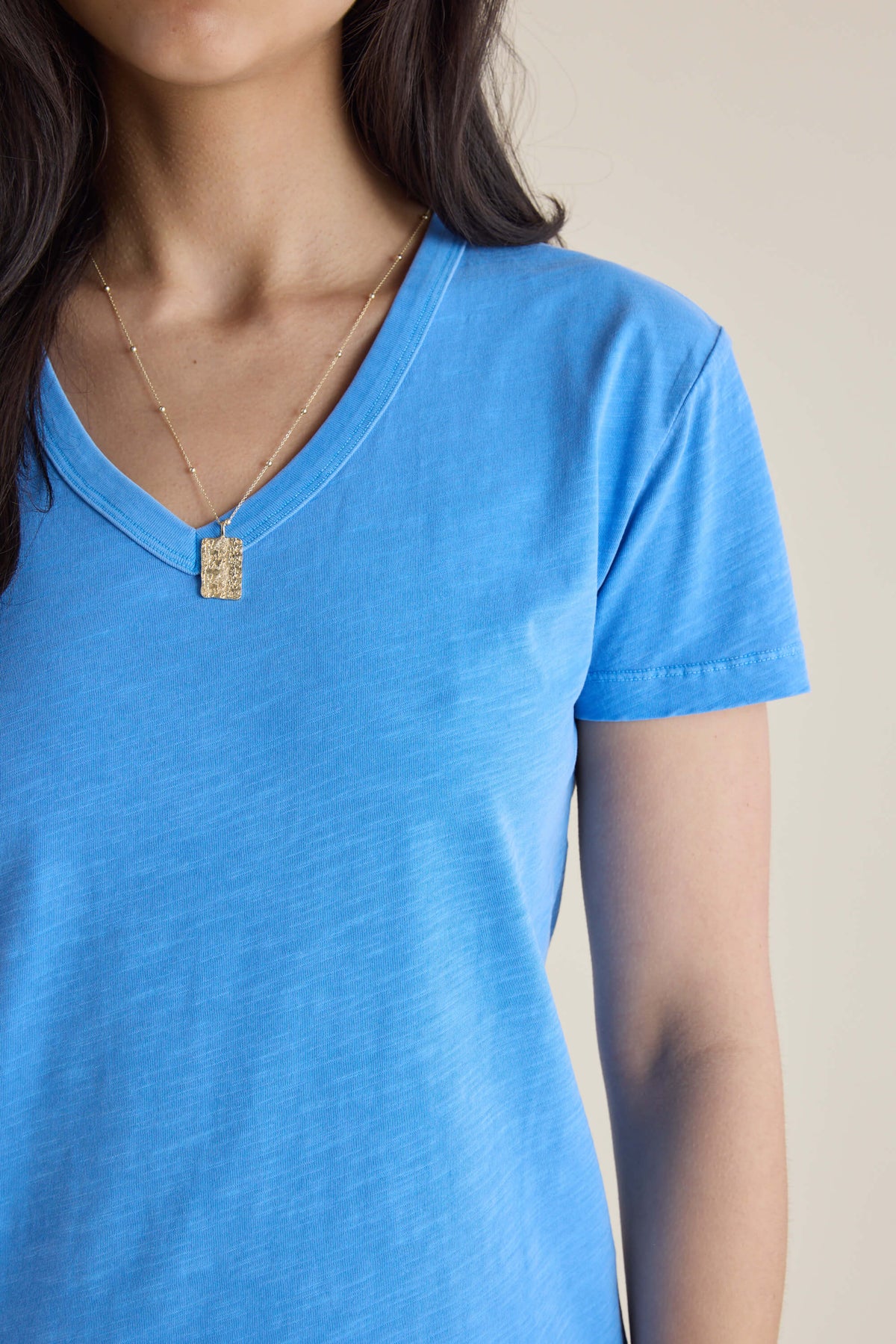 Woman in blue Elementary V Neck Tee, organic cotton, gold necklace; shoulders to chest shown.