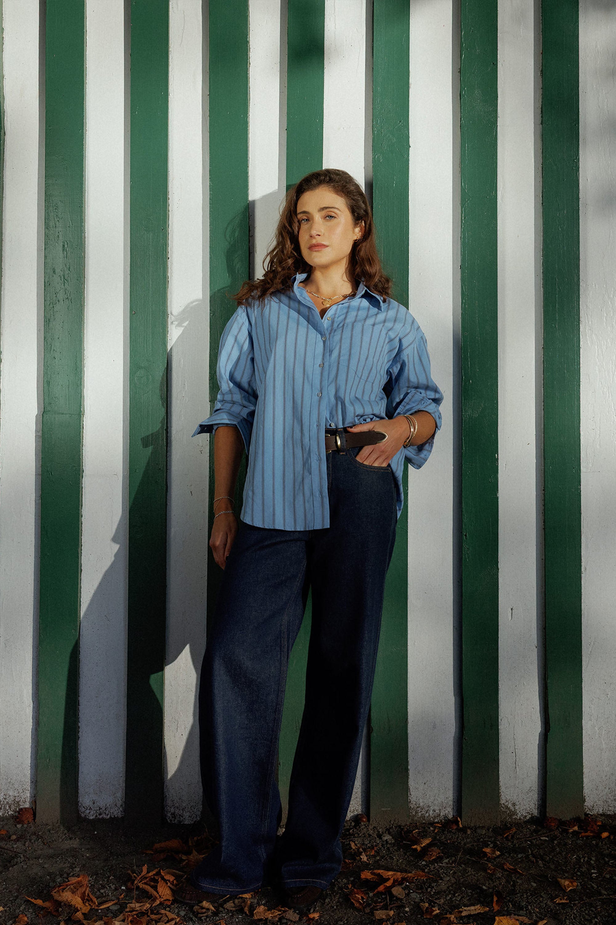 Woman in Nevada Wide Jeans by Max and blue shirt stands by a green and white striped wall.