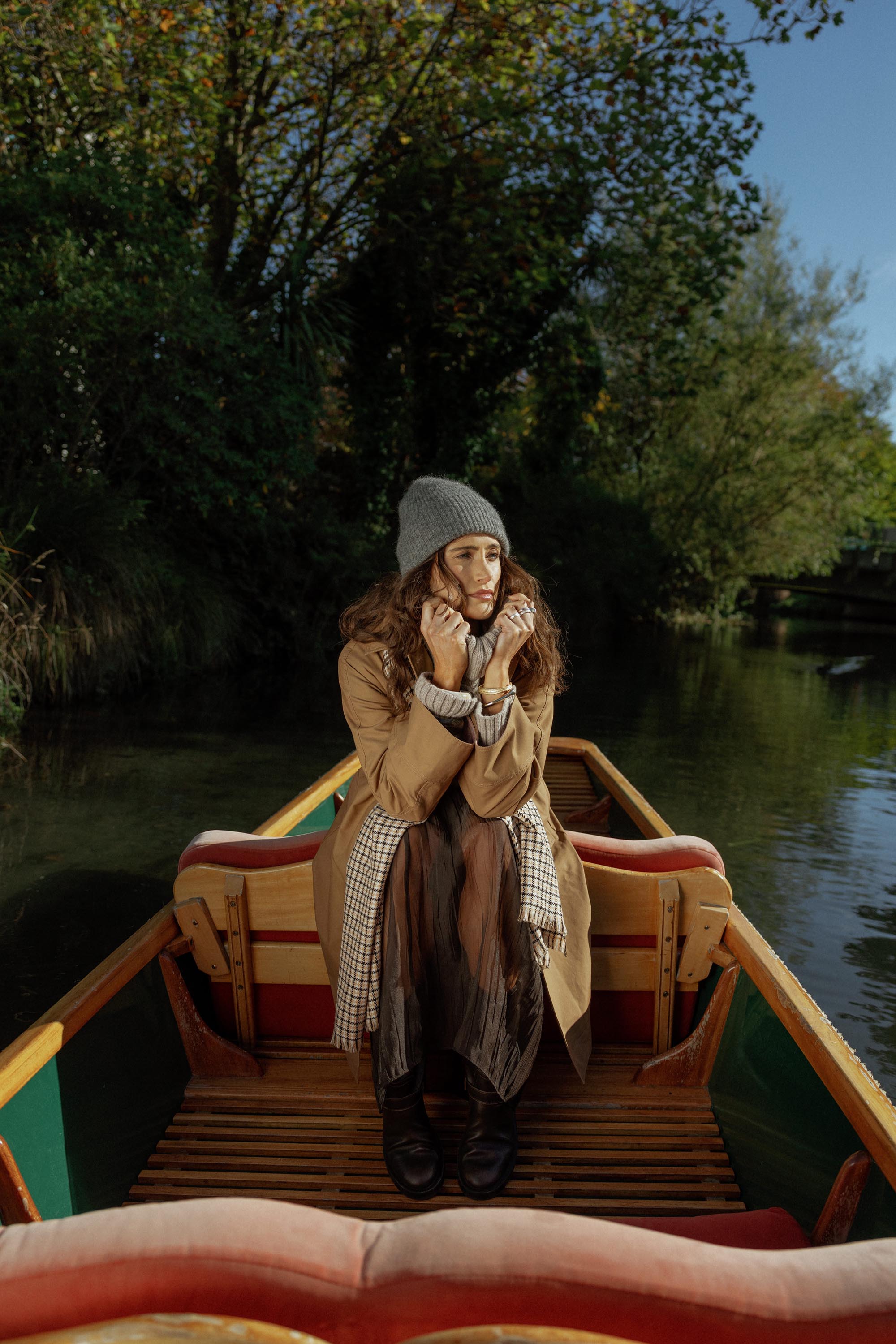 A woman in a Max London Trench Coat and gray beanie sits on a boat on a calm river.