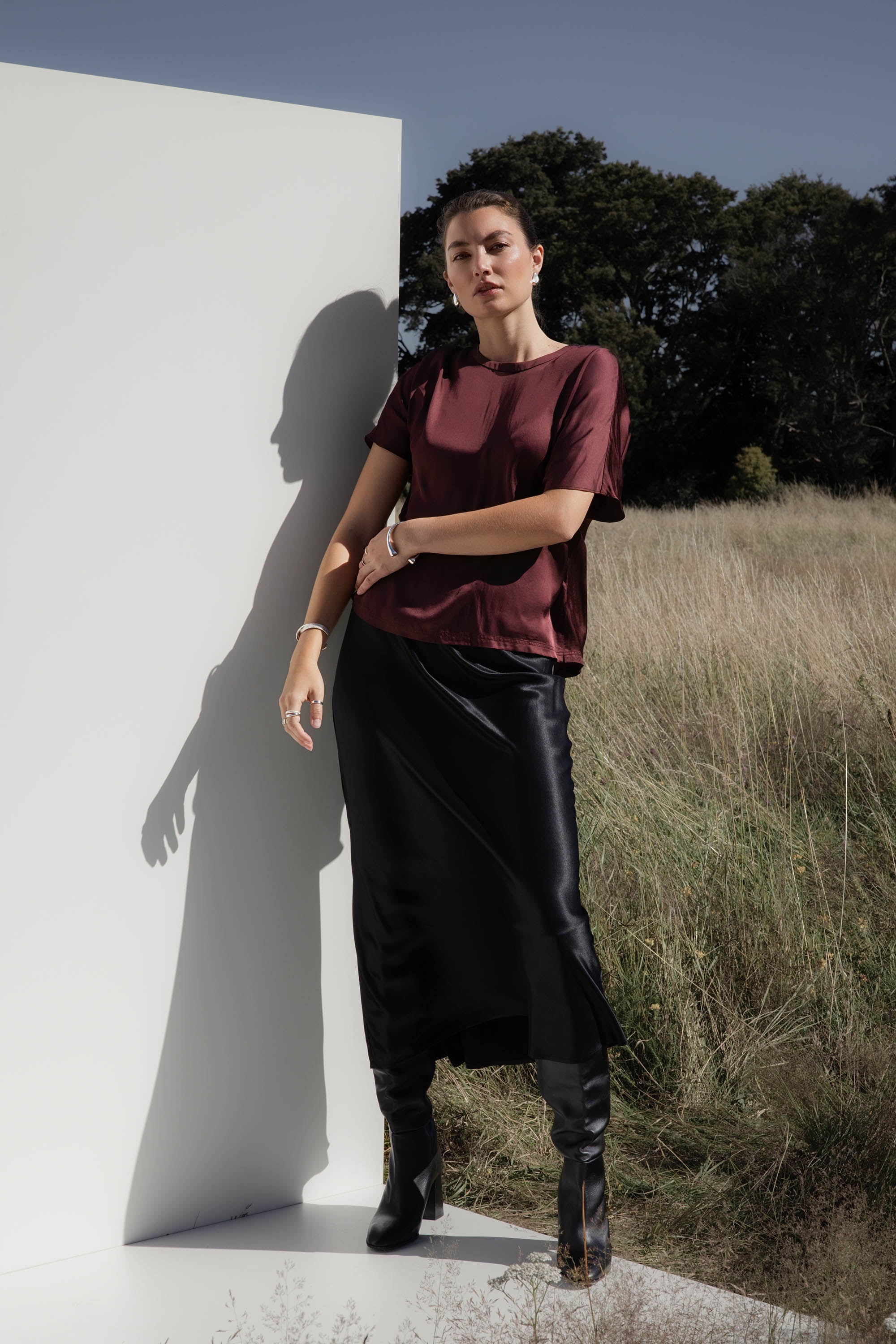 Woman outdoors wears Max’s The Perfect Slip Skirt with a maroon top by a white wall in tall grass.
