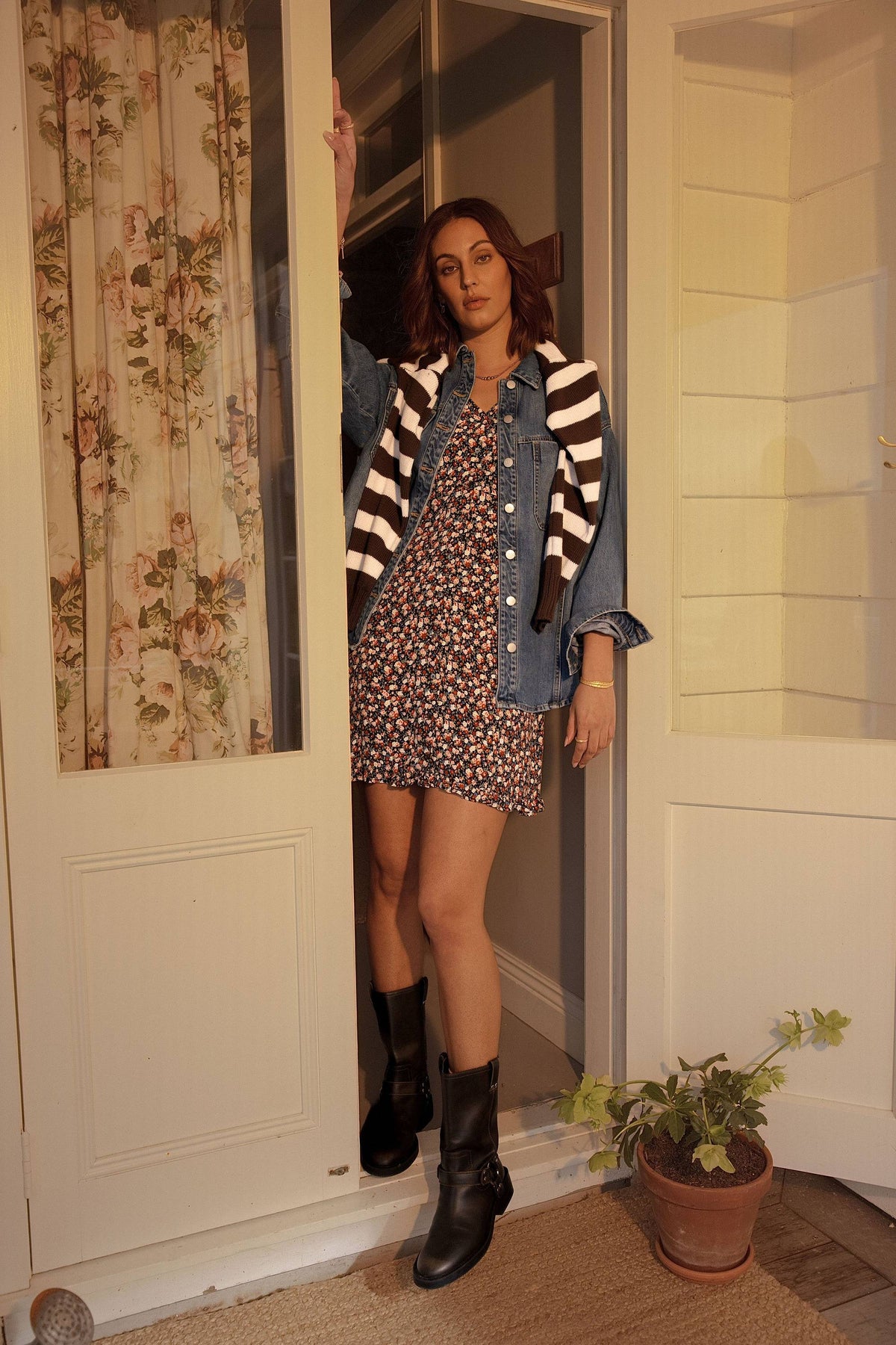 Woman in Elementary Button Front Dress and boots stands in doorway by a potted plant.