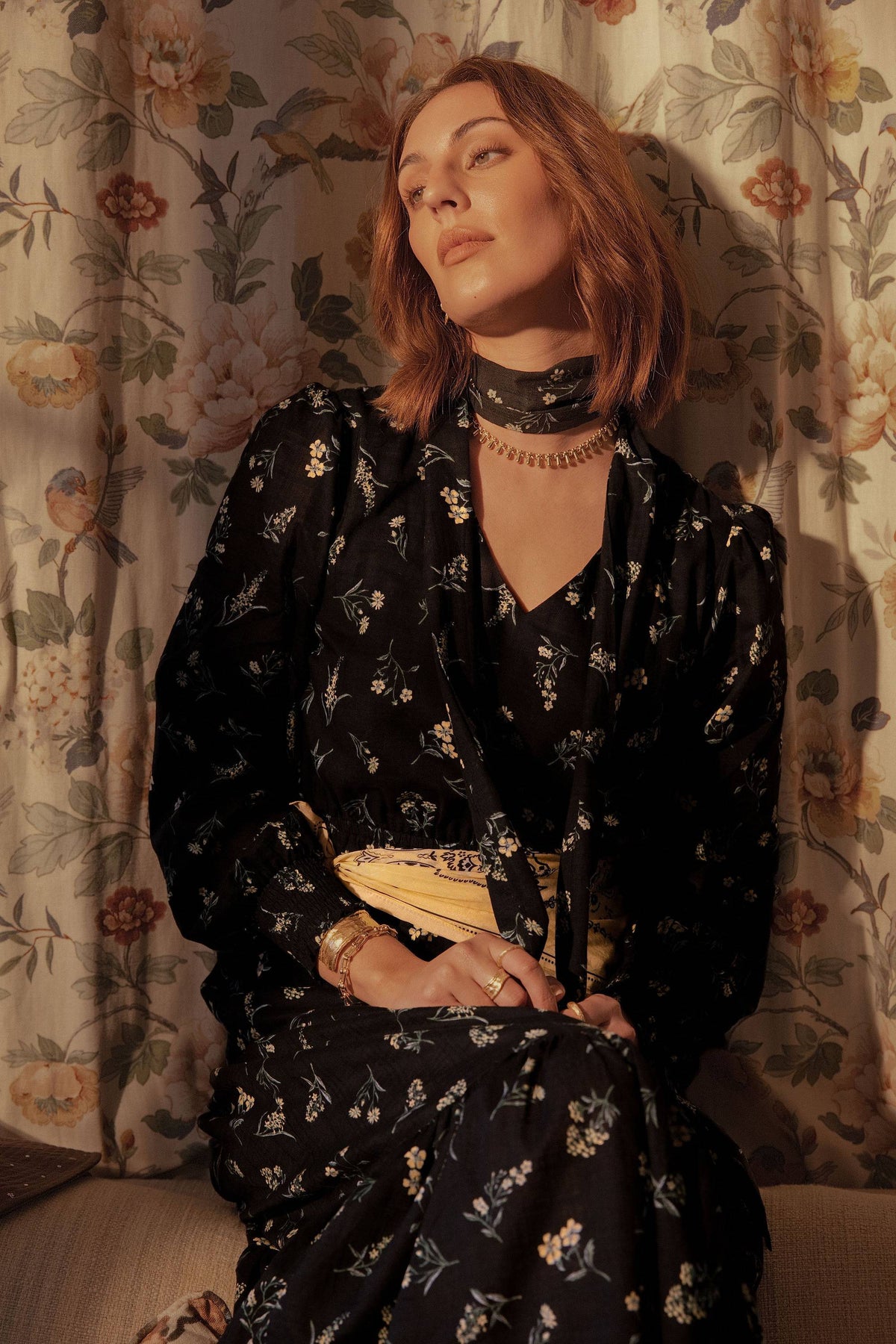 Woman in a black Elementary Tiered Dress by Elementary sits on a couch, gazing to the side.