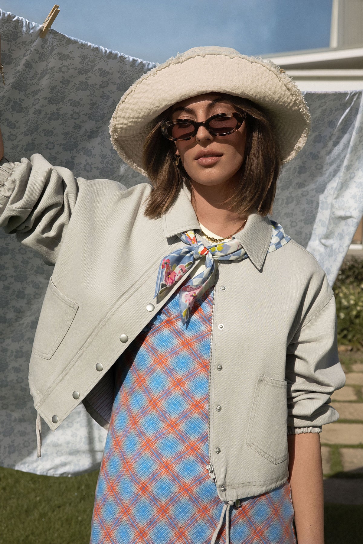 A woman standing in front of a washing line wearing a blue checkered dress, a grey jacket and a bucket hat.