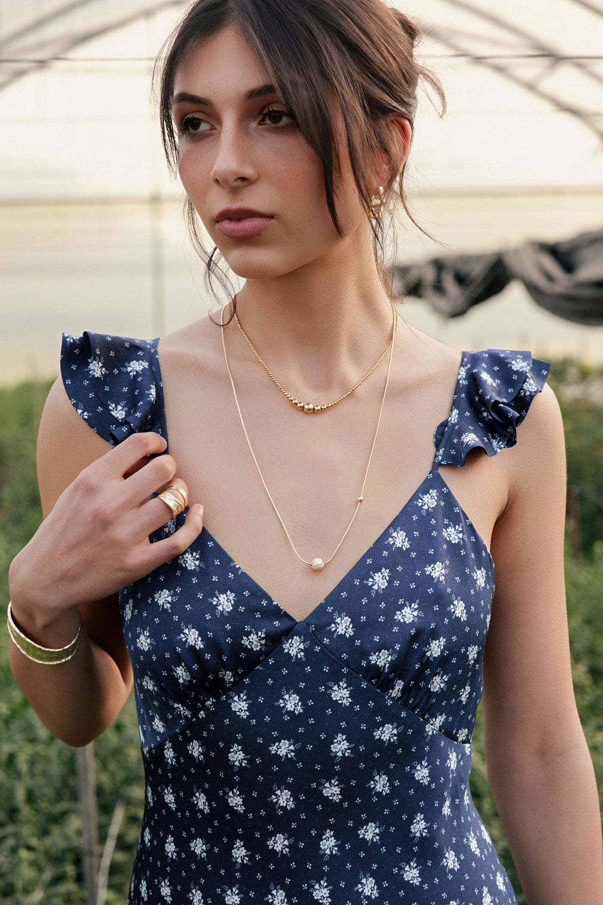 A woman standing in a greenhouse wearing a blue floral dress and gold jewellery.