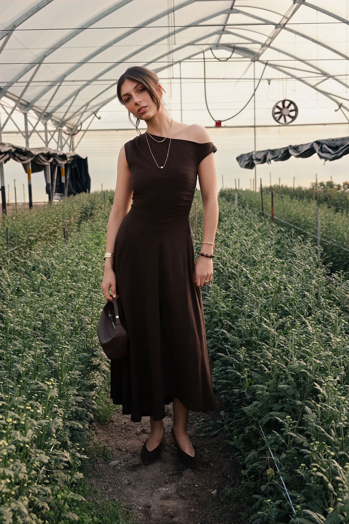 A woman standing in a greenhouse wearing a brown dress.