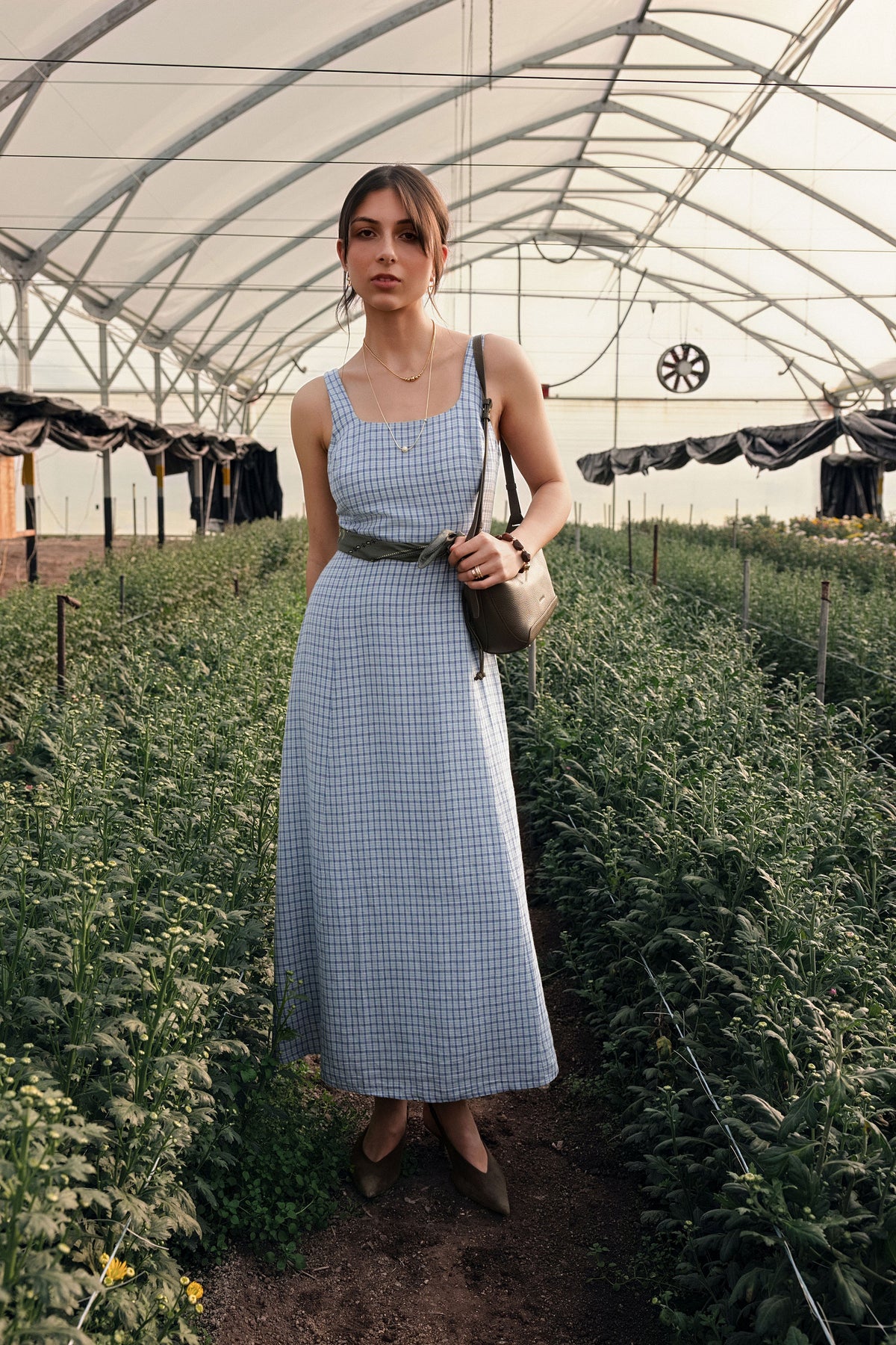 A woman standing in a greenhouse wearing a blue checkered dress.