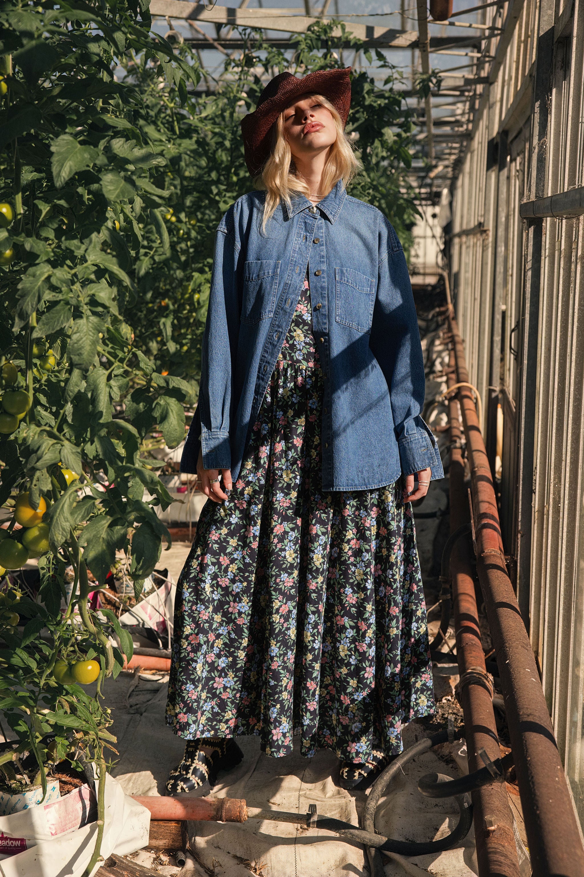 A woman standing in a greenhouse wearing a floral spring midi dress layered with a denim shirt.