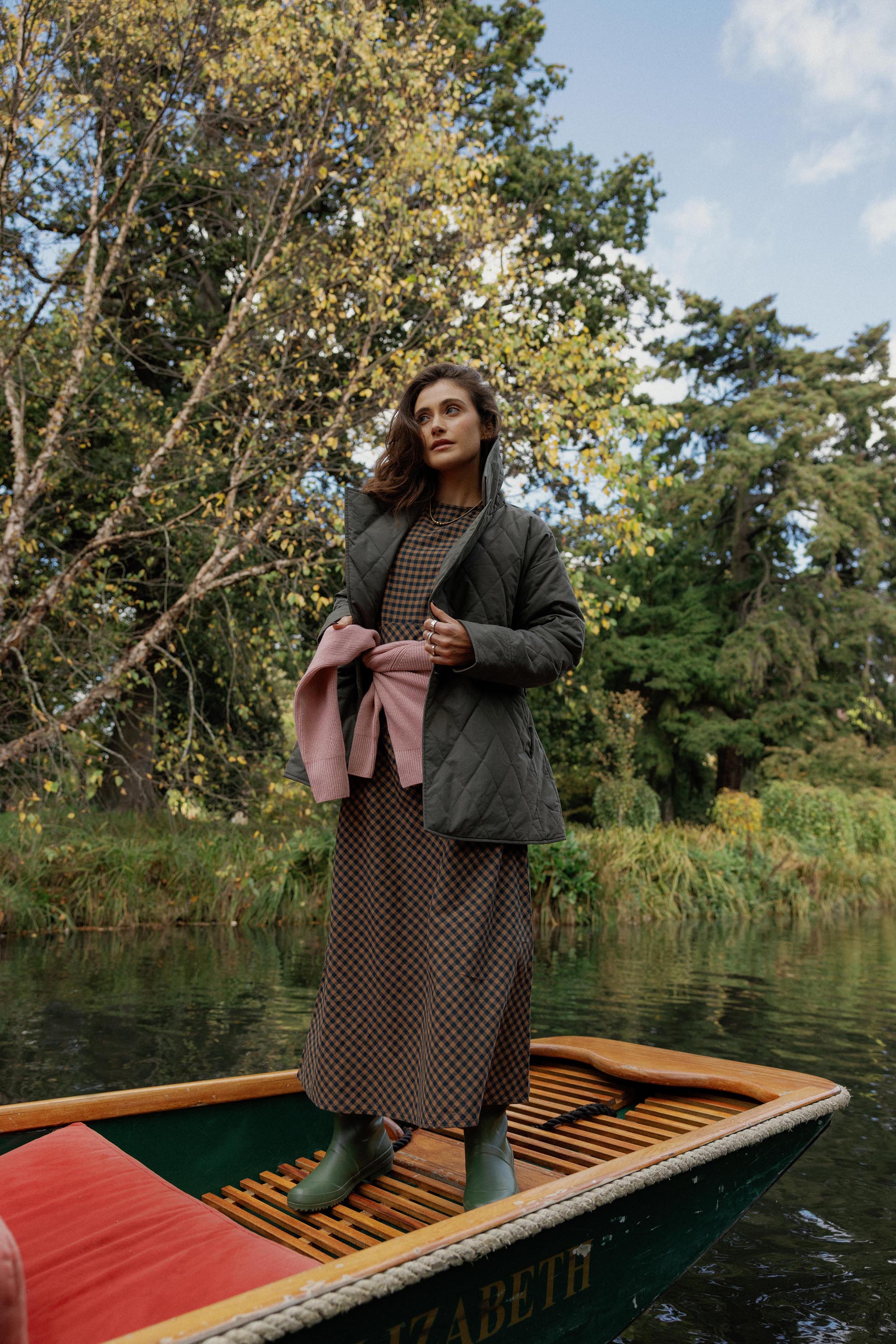 A woman on a wooden boat wears the Max Lucy Bias Cut Dress and boots among riverside trees.