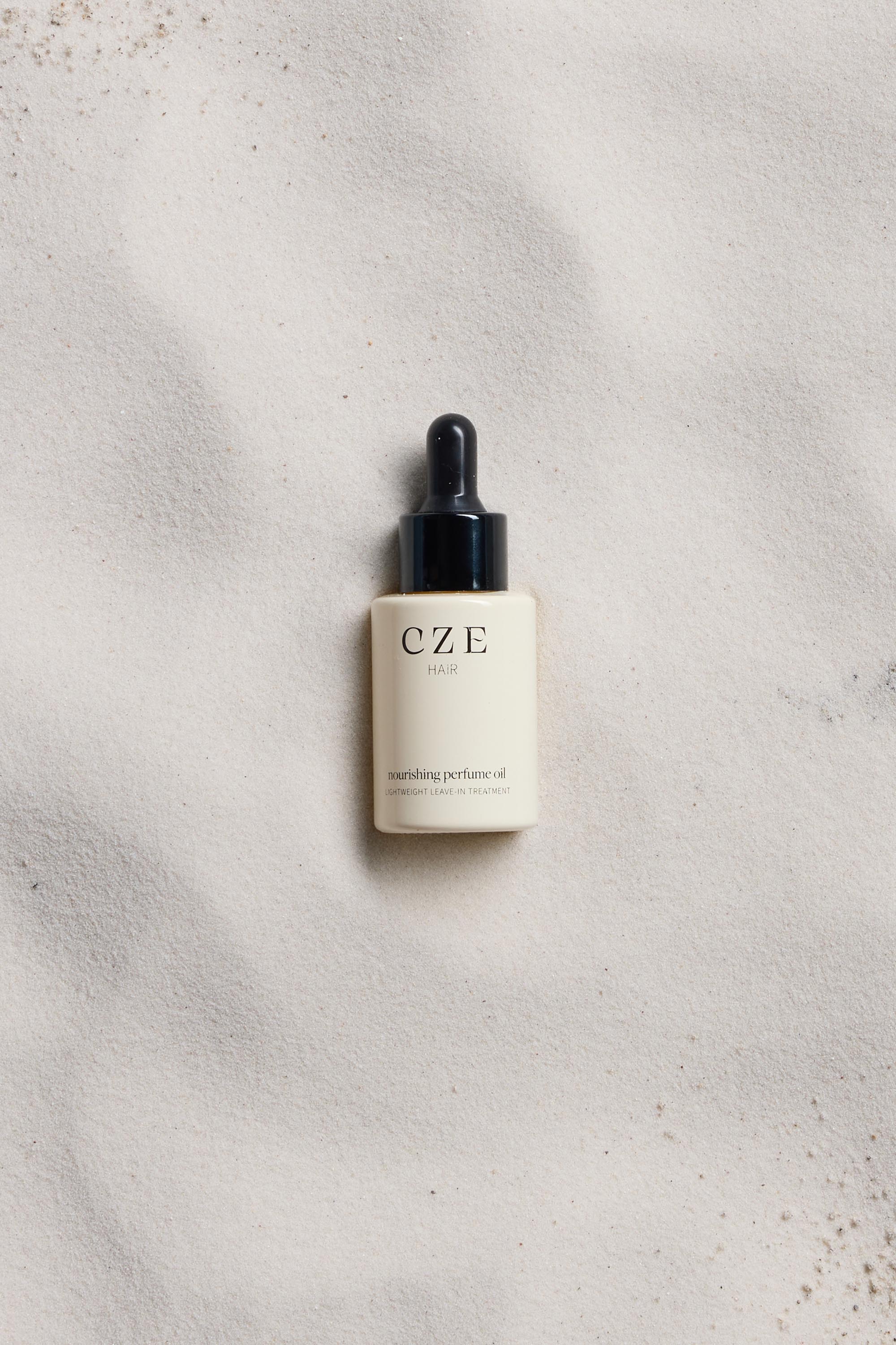 A 30ml CZE Hair Nourishing Perfume Oil dropper bottle by CZE Hair, perfect as a pre-styling oil to control frizz, is placed on a smooth white sandy surface.