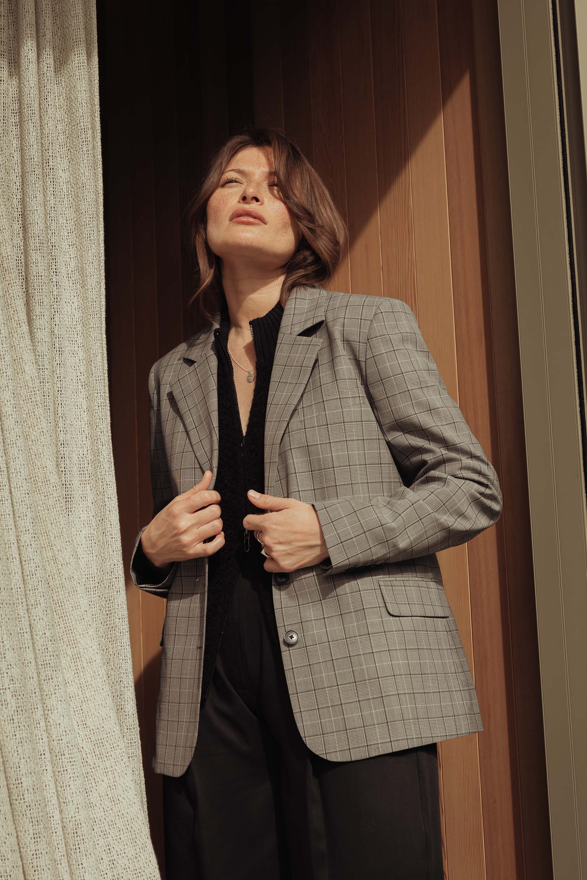 A person with short brown hair stands by a sunlit window, adjusting The Wool Blend Blazer by Max over a black outfit. Warm light highlights their face and the wooden wall behind them.