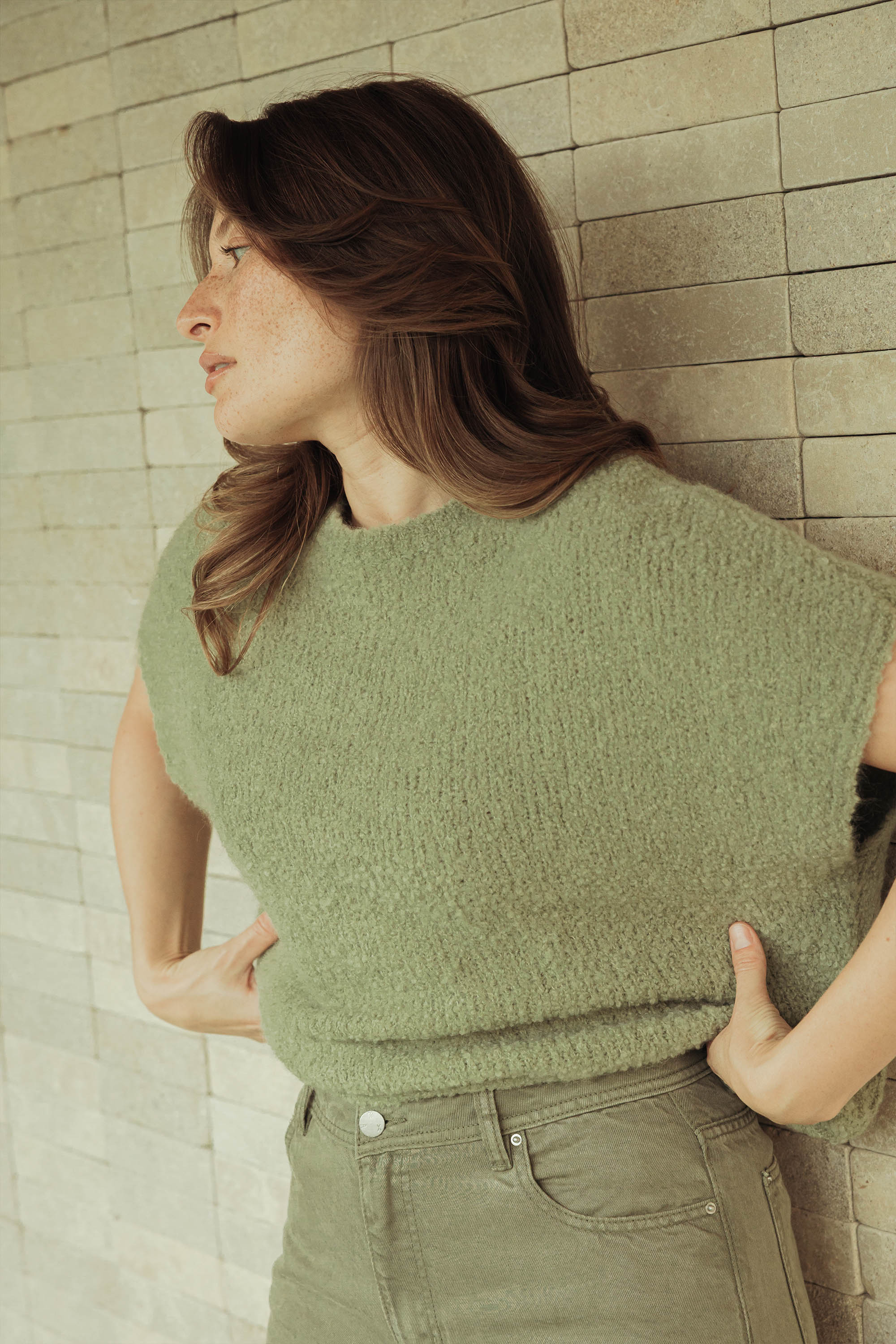 A woman with light brown hair and freckles wears the sage green Elementary Short Sleeve Top by Elementary and green pants, standing against a tiled beige wall, looking sideways with her hands on her hips.