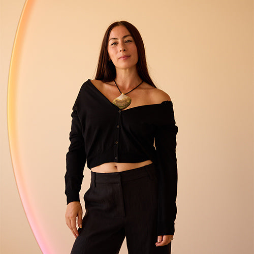 A woman with long brown hair wears a black off-the-shoulder top, black pants, and a large stone pendant necklace. She stands against a beige background with a soft, curved rainbow light effect on the left side.