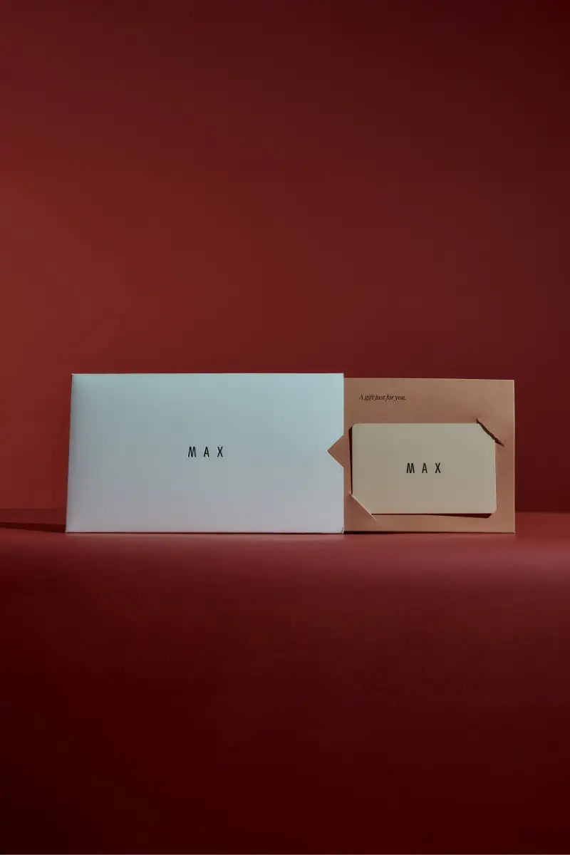 A white envelope marked Max and a beige card holder, both with the Max logo, sit on a rich reddish-brown background—ideal for presenting Max Digital Gift Cards with a personal message.